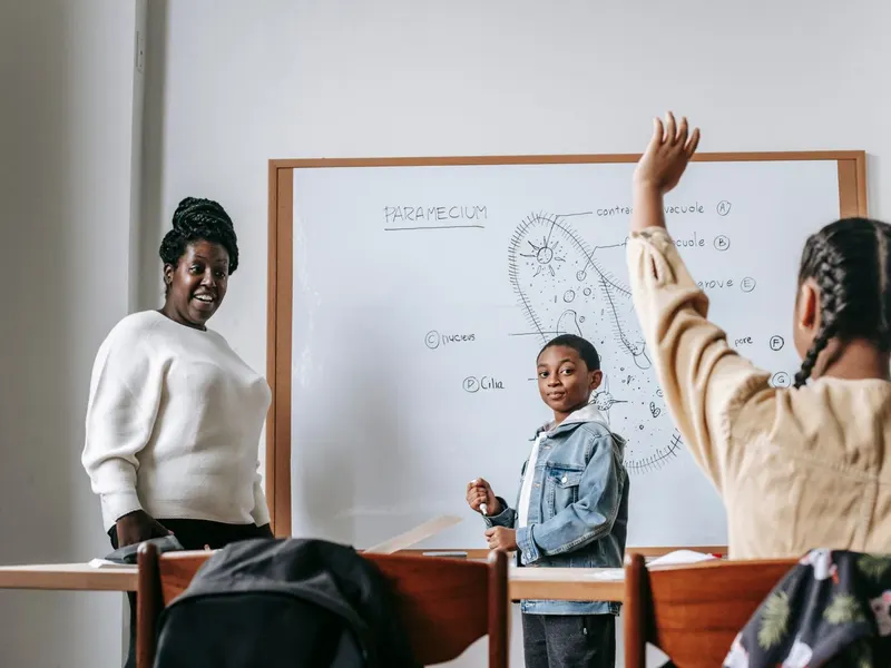 A smiling teacher standing beside a whiteboard while pupils raise their hands in class